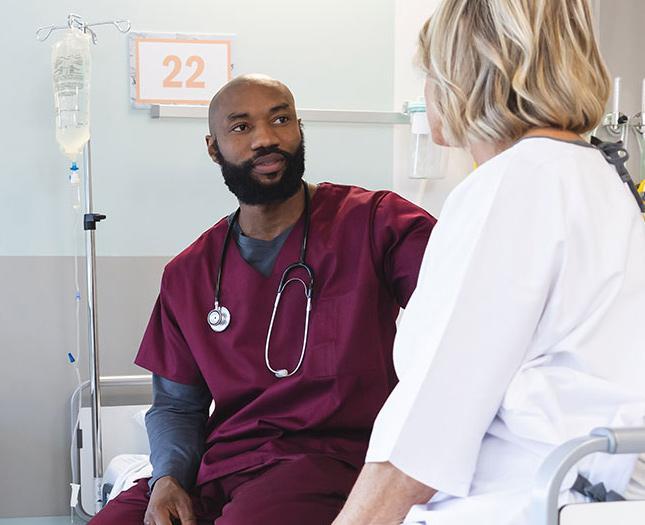 Join our team Staff member giving eye contact to a patient while they both sit on a hospital bed.
